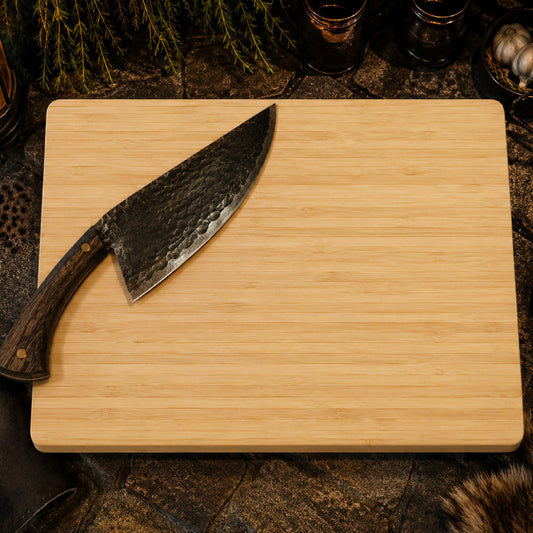 A rectangular light wood cutting board with a large chef's knife resting on the top-left corner, set against a rustic dark stone and wood background with herbs and glassware.