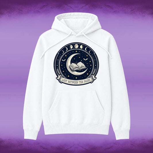 A white pullover hoodie with a circular celestial graphic on the chest featuring a crescent moon, open book, stars, and moon phases, with the text 'LOST BETWEEN THE PAGES' on a banner. The hoodie is shown against a purple textured background.