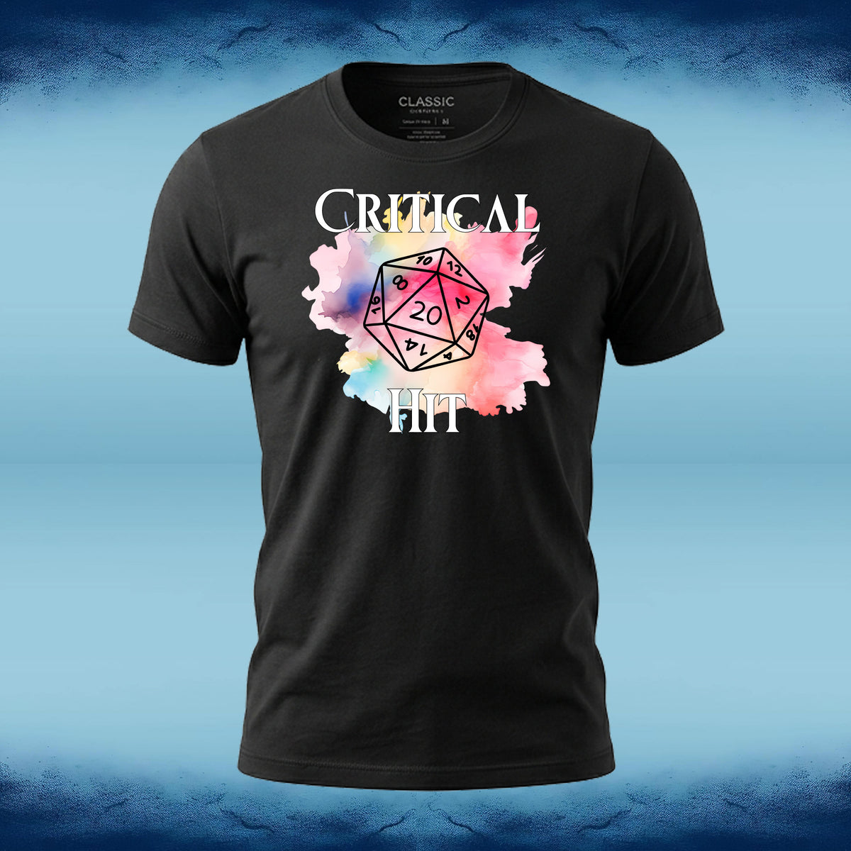 A black short-sleeve t-shirt with a graphic print featuring the word 'CRITICAL' in white, a pink and blue watercolor splatter, a black outlined 20-sided die with 'D20' in the center, and 'IT IS' below. The shirt is displayed against a blue textured background.
