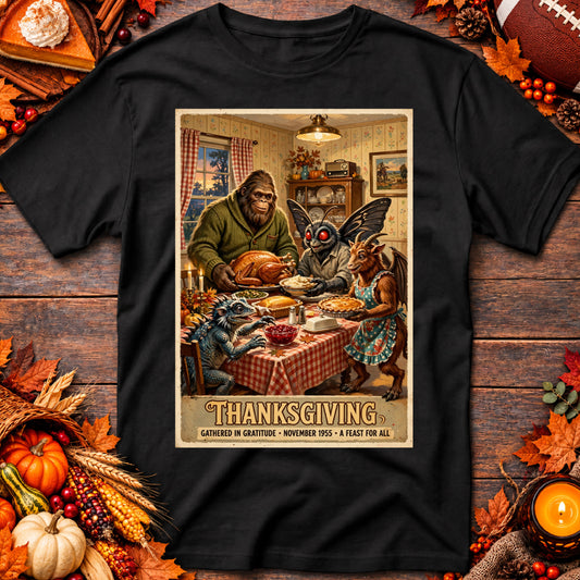 Black graphic tee featuring Bigfoot, Mothman, and fellow cryptids sharing a Thanksgiving feast, laid flat among autumn decorations.