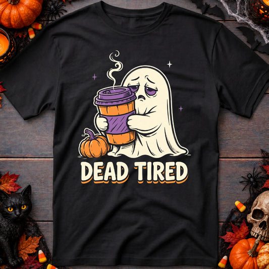 A black short-sleeve graphic t-shirt laid flat on a dark wood background with Halloween decorations. The graphic shows a sad white ghost holding a purple and orange coffee cup, with the text 'DEAD TIRED' below it.