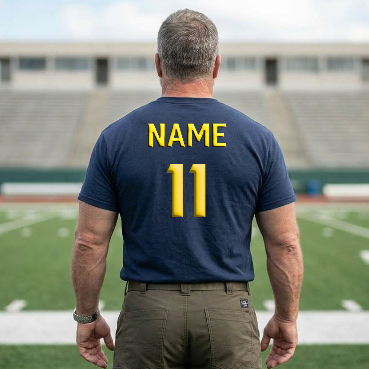 A man viewed from behind wearing a navy blue t-shirt with 'NAME 11' printed in yellow on the back, standing on a football field with stadium seating in the background.