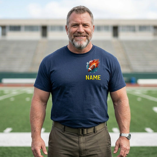 A smiling man with a gray beard stands on a football field wearing a navy blue t-shirt with a custom graphic of a flaming football and the text 'NAME' in yellow. The background shows blurred stadium seating and the field.