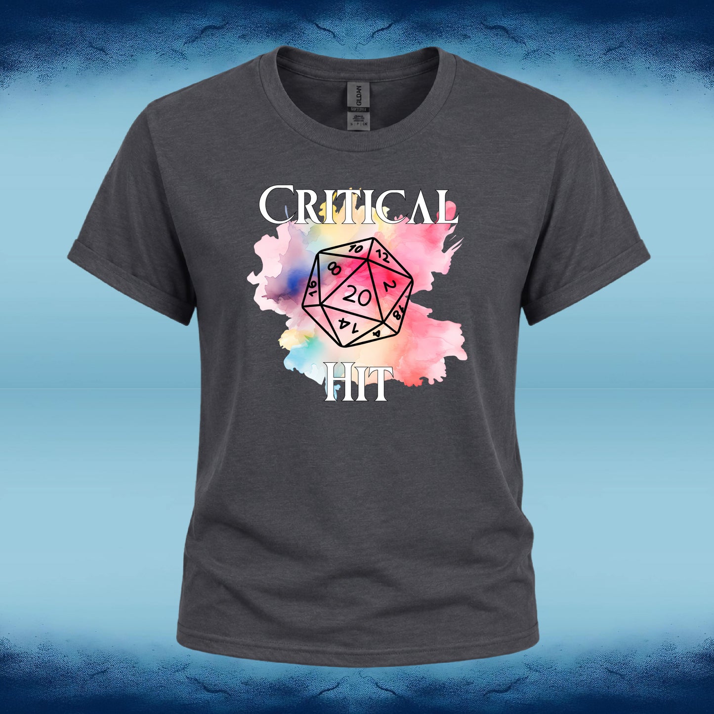 A dark gray crewneck t-shirt featuring a graphic print of a 20-sided die over a pink and blue watercolor splash, with the words 'CRITICAL HIT' in white text. The shirt is displayed against a blue textured background.