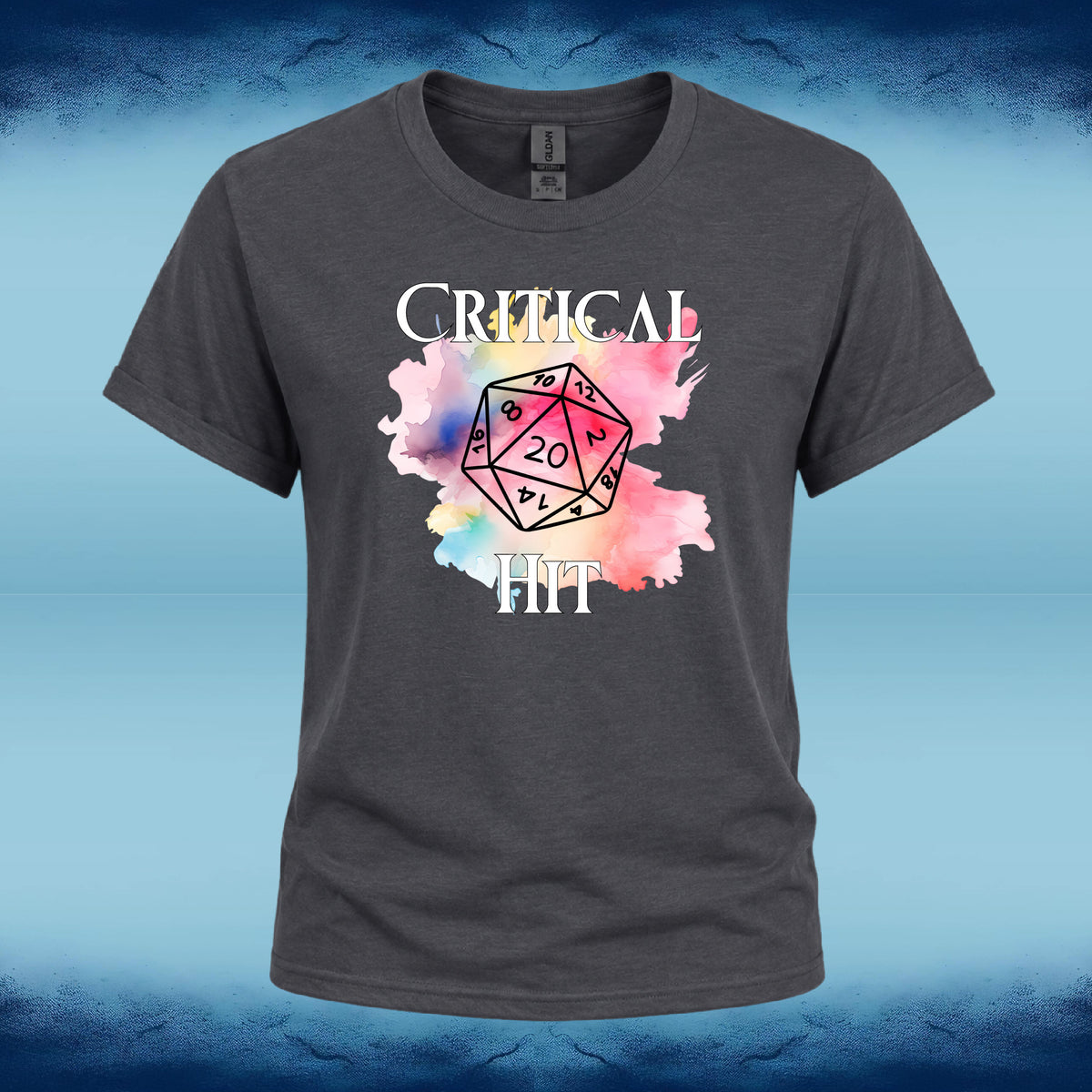 A dark gray crewneck t-shirt featuring a graphic print of a 20-sided die over a pink and blue watercolor splash, with the words 'CRITICAL HIT' in white text. The shirt is displayed against a blue textured background.