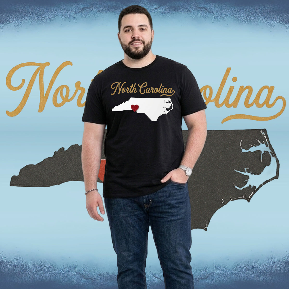 A man wearing a black t-shirt with a white North Carolina state graphic and a small red heart, standing in front of a blue graphic background with gold 'North Carolina' script and a dark state silhouette.