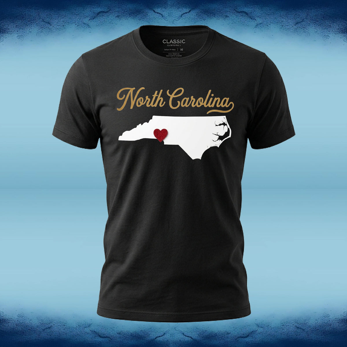 A black short-sleeve t-shirt with a graphic of the white silhouette of North Carolina, featuring a small red heart in the middle and 'North Carolina' written in gold script above it. The shirt is displayed against a blue textured background.