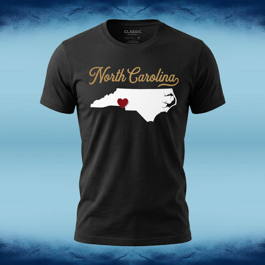 A black short-sleeve t-shirt with a graphic of the white silhouette of North Carolina, featuring a small red heart in the middle and 'North Carolina' written in gold script above it. The shirt is displayed against a blue textured background.