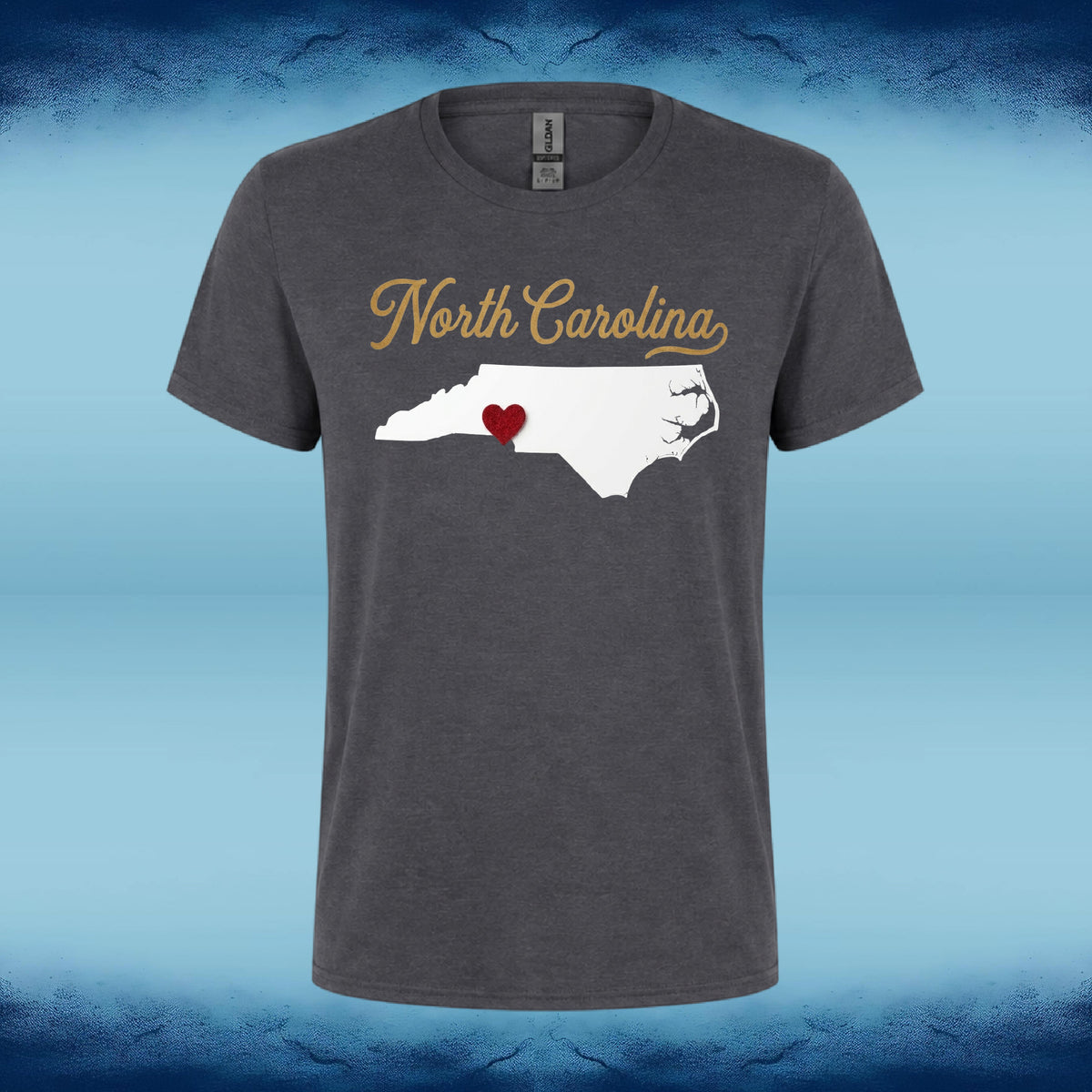 A dark heather gray t-shirt featuring a graphic of the state map of North Carolina with the words 'North Carolina' in gold script and a small red heart highlighting the boundary between North and South Carolina. The shirt is displayed against a blue gradient background with a textured dark blue border.