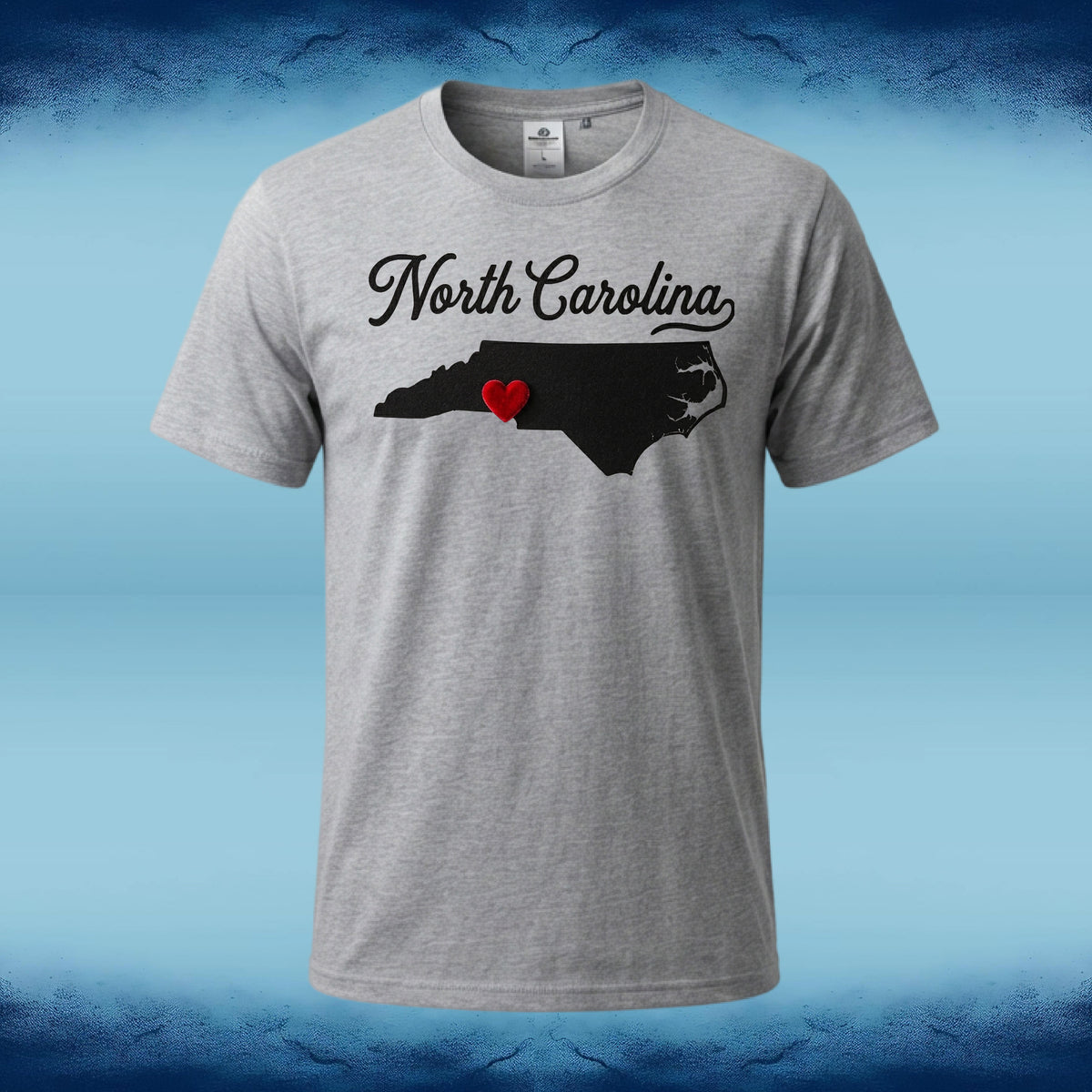 A heather gray short-sleeve t-shirt featuring a black graphic of the state map of North Carolina with a small red heart in the center and 'North Carolina' written in black script above it. The t-shirt is displayed against a blue gradient background with a dark blue textured border.