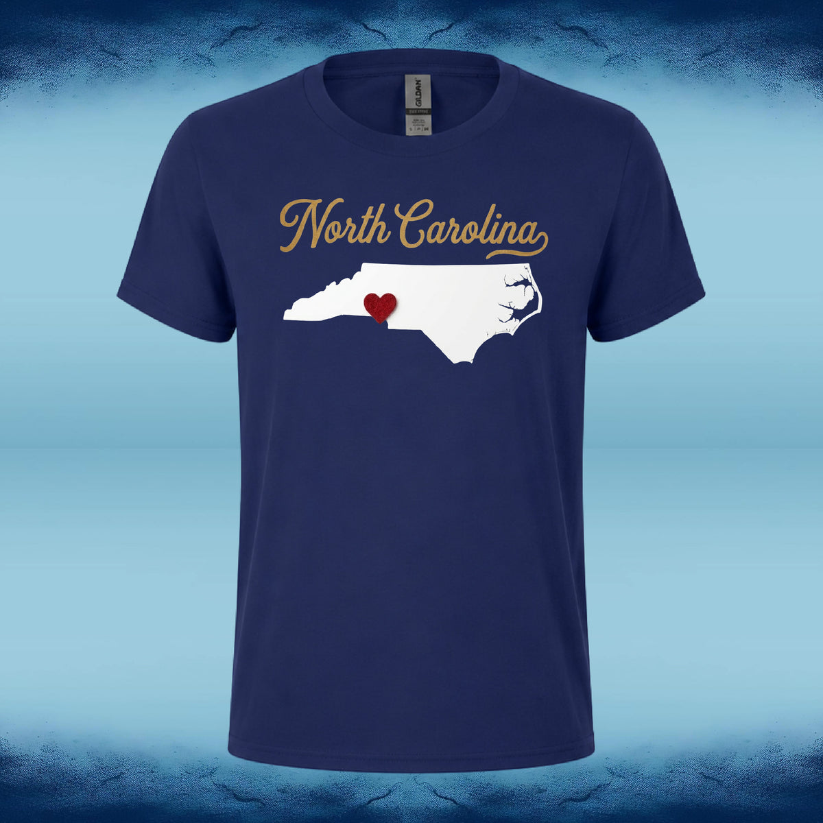 A navy blue short-sleeve t-shirt with a graphic on the chest featuring the words 'North Carolina' in gold script, a white map outline of North Carolina, and a small red heart inside the state. The shirt is displayed against a blue textured background.