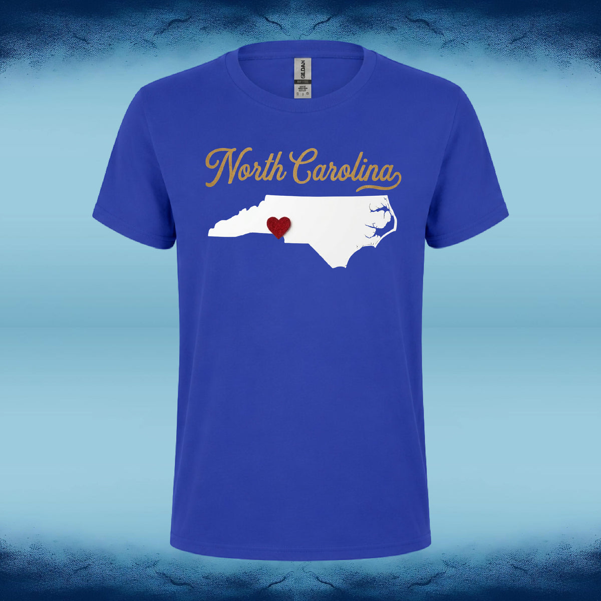 A royal blue short-sleeve t-shirt with a graphic on the chest featuring the words 'North Carolina' in gold cursive and a white silhouette of North Carolina with a small red heart inside, shown against a blue gradient background.
