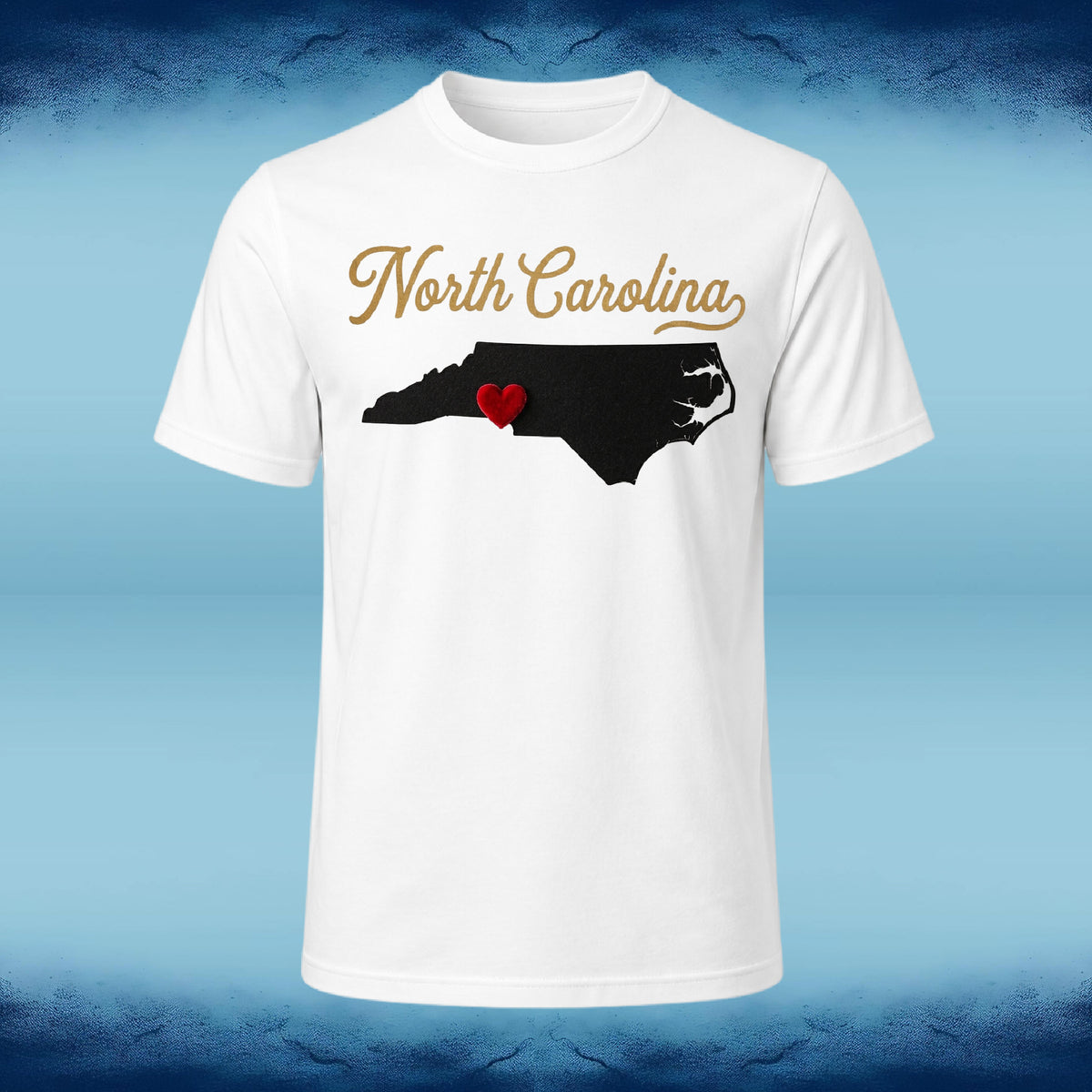 A white short-sleeve crewneck t-shirt featuring a graphic of the state map of North Carolina in black with a small red heart, and the words 'North Carolina' in gold script above it, displayed against a blue textured background.