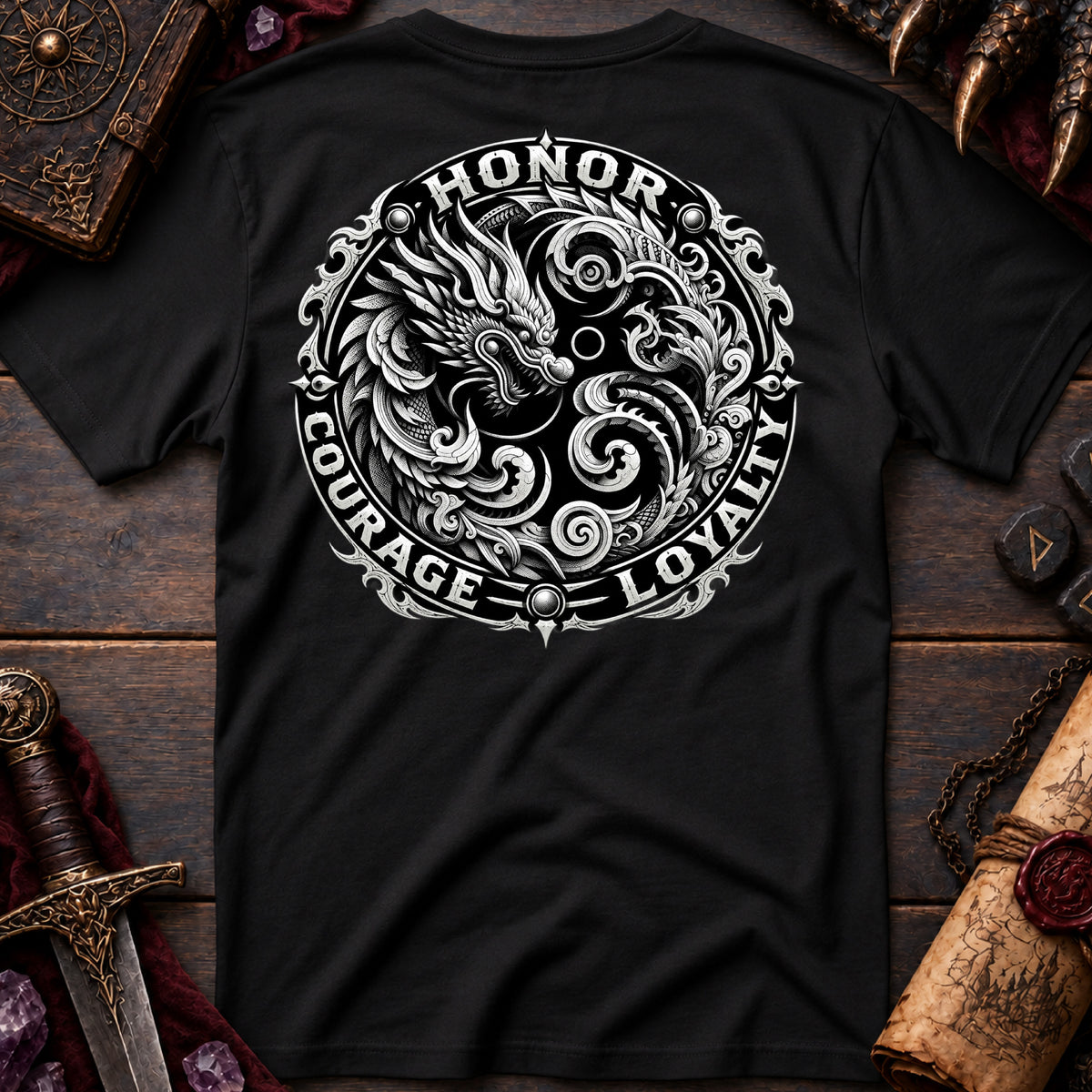 A black short-sleeve T-shirt laid flat on a dark wooden surface, featuring a large circular white graphic on the back with a dragon illustration and the words HONOR, COURAGE, and LOYALTY. Decorative props including a sword, scroll, crystals, and leather-bound book surround the shirt.