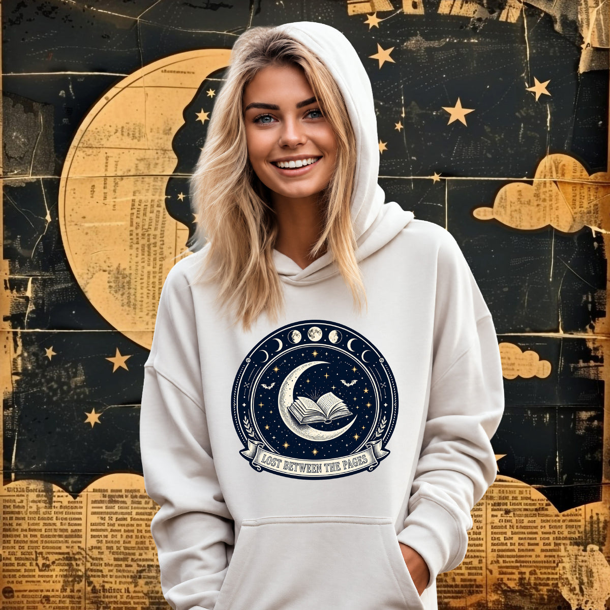 A smiling woman with blonde hair wears a White-colored hoodie featuring a dark blue circular graphic with moon phases, an open book, stars, and the text 'LOST BETWEEN THE PAGES'. The background is a textured collage of a gold moon, stars, and book pages.