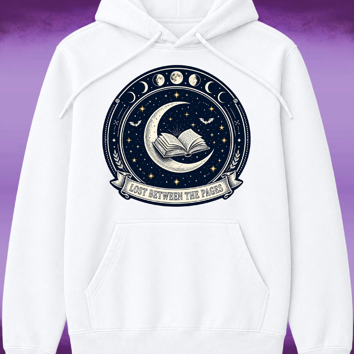 A white pullover hoodie with a large circular celestial graphic on the chest. The graphic features a dark navy blue night sky with a crescent moon, an open book, stars, moon phases, bats, and a banner reading 'LOST BETWEEN THE PAGES'.