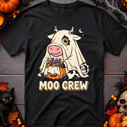 A black short-sleeve graphic T-shirt laid flat on a dark wood surface, surrounded by Halloween props including pumpkins, a black cat figurine, spiderwebs, and a skull. The shirt features a cartoon cow in a white ghost costume holding a pumpkin with purple grapes, above the words 'MOO CREW'.