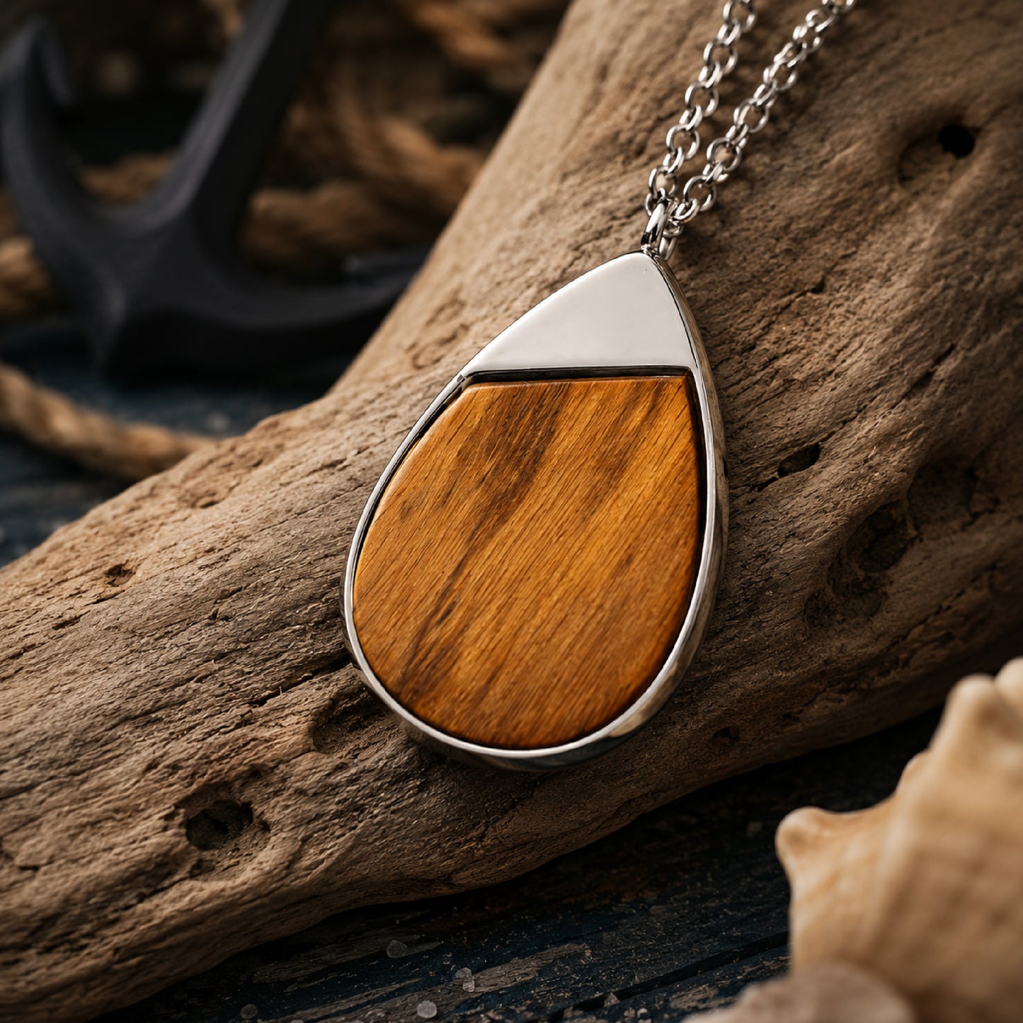 A teardrop-shaped pendant necklace with a polished silver-tone metal frame and a warm brown wood inlay, resting on a piece of driftwood with a nautical anchor and rope in the background.