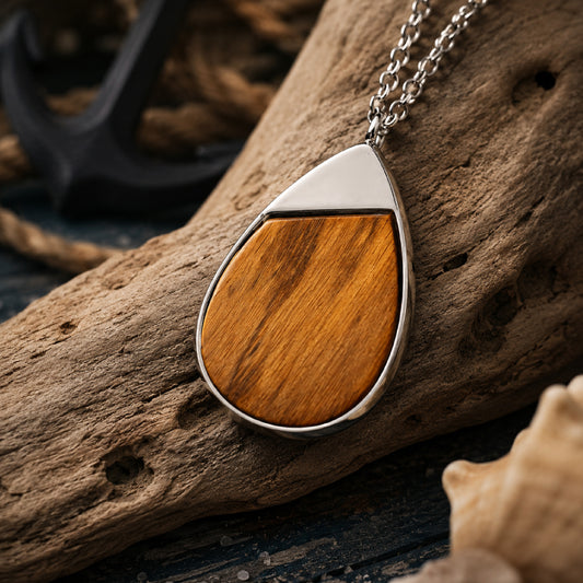 A teardrop-shaped pendant necklace with a polished silver-tone metal frame and a warm brown wood inlay, resting on a piece of driftwood with a nautical anchor and rope in the background.