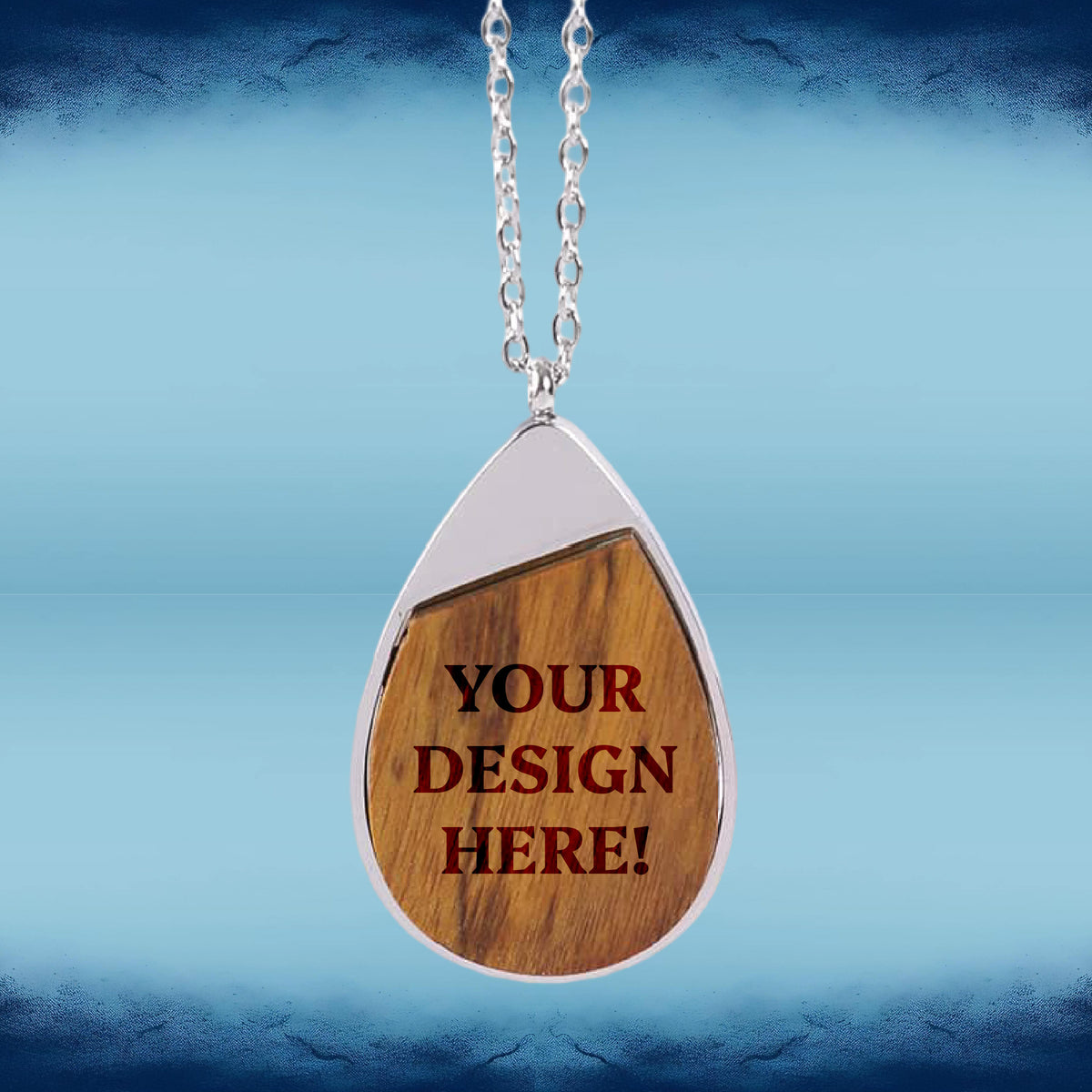 A teardrop-shaped pendant necklace with a silver-tone chain and border, featuring a wood-grain center with the text 'YOUR DESIGN HERE!' in red, shown against a blue gradient background.