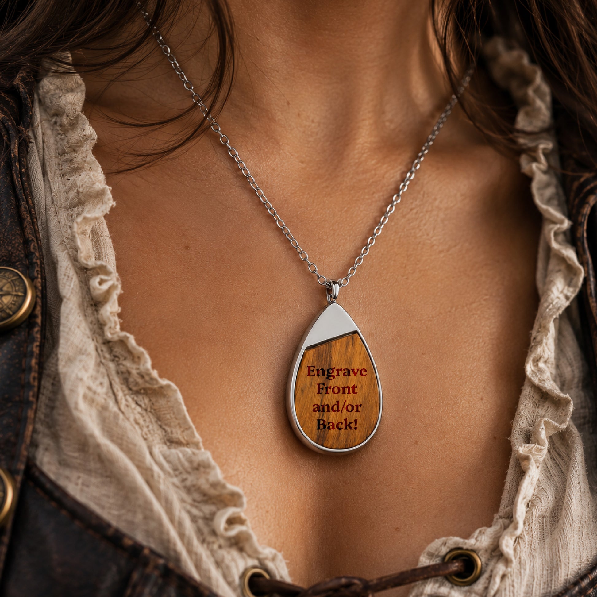 A close-up of a person wearing a silver teardrop pendant necklace with a wood inlay engraved with the text 'Engrave Front and/or Back!'. The person is wearing a ruffled cream blouse and a dark leather jacket.