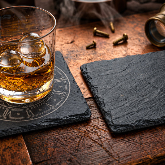 Two black slate coasters on a rustic wooden table, with one coaster holding a glass of amber whiskey and ice and the other left empty.