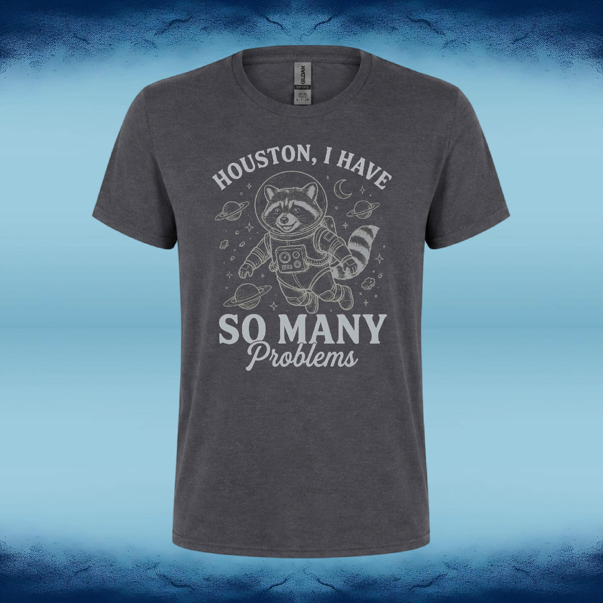 A dark heather gray t-shirt featuring a distressed white graphic of a raccoon in a space suit with the text 'HOUSTON, I HAVE SO MANY Problems'. The shirt is displayed against a blue gradient background with a textured dark blue border.