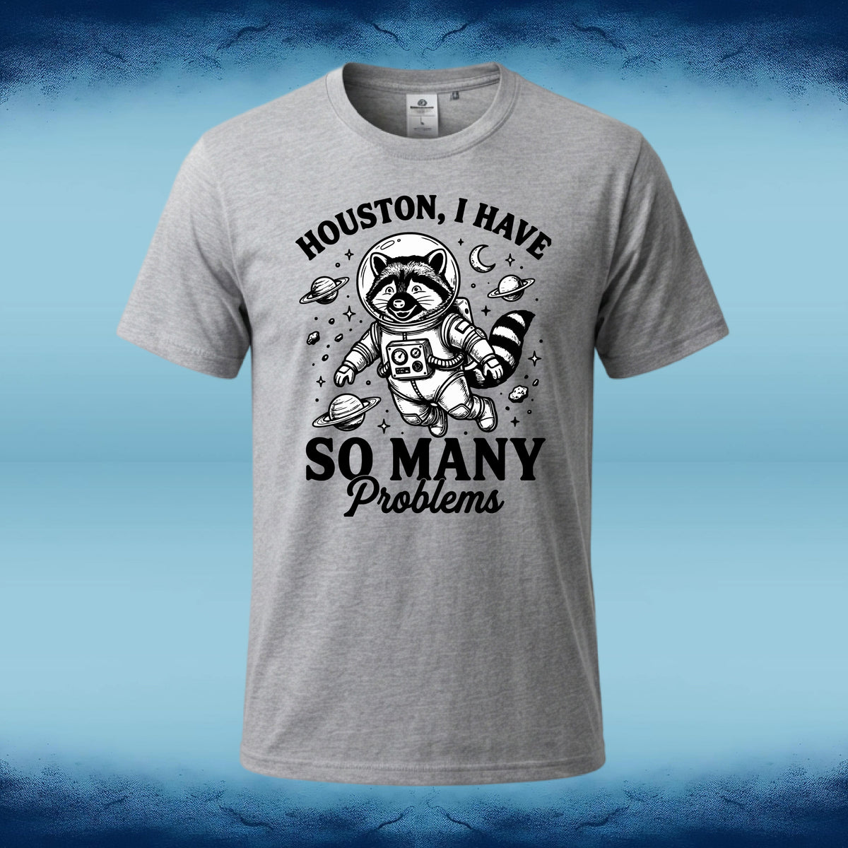 A heather gray short-sleeve t-shirt with a black graphic of a raccoon astronaut floating in space with planets and the text 'HOUSTON, I HAVE SO MANY Problems', shown against a blue gradient background.
