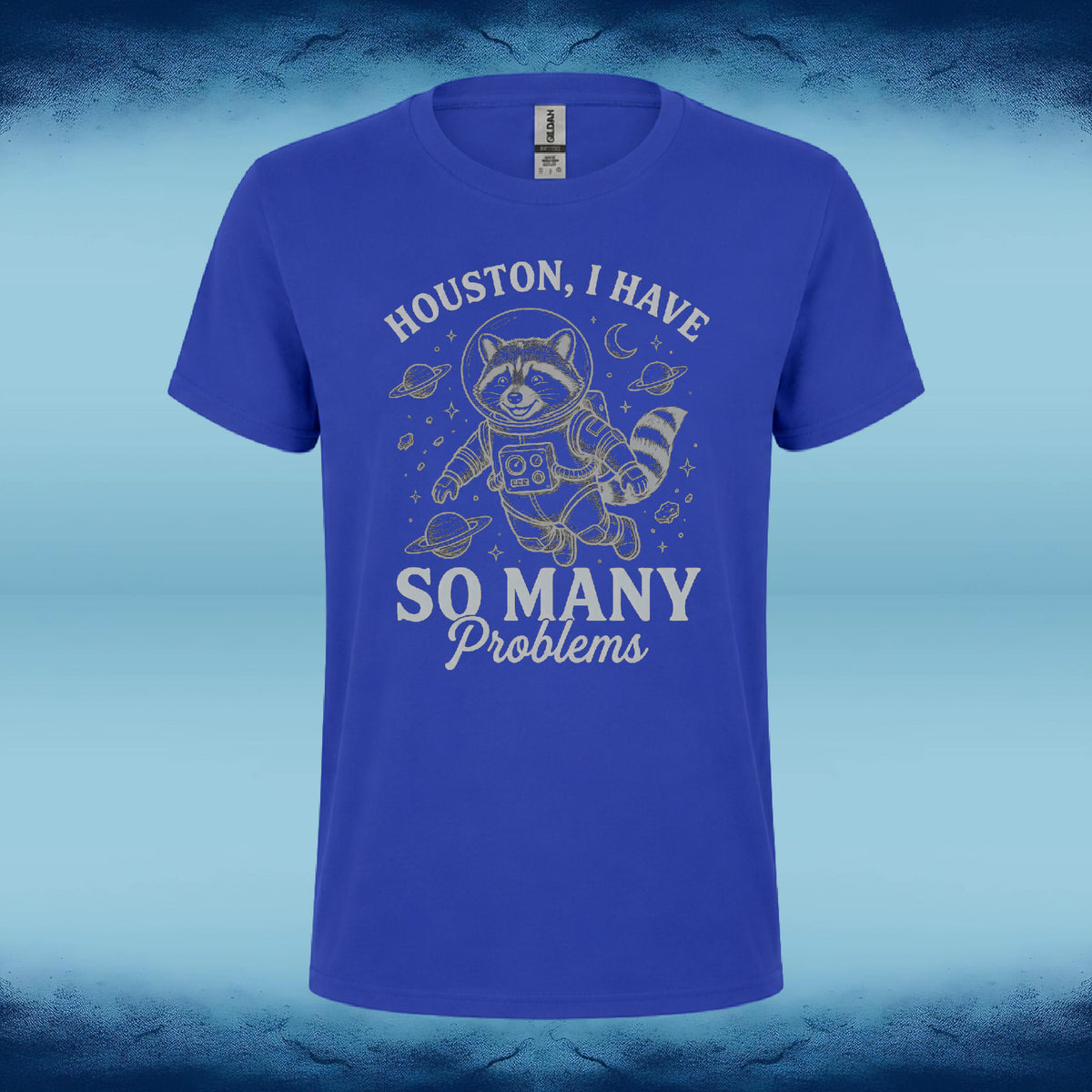 A royal blue short-sleeve t-shirt with a graphic of a cartoon raccoon astronaut floating in space, with the text 'HOUSTON, I HAVE SO MANY Problems' printed in white. The shirt is displayed against a blue and black textured background.